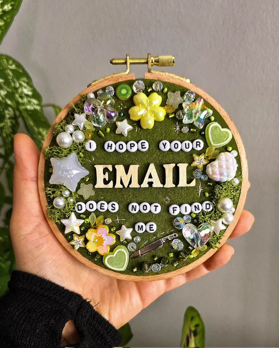 A hand holding an ornate bead embroidery hoop, with various beads, sequins and moss. The beads read "I hope your email does not find me"