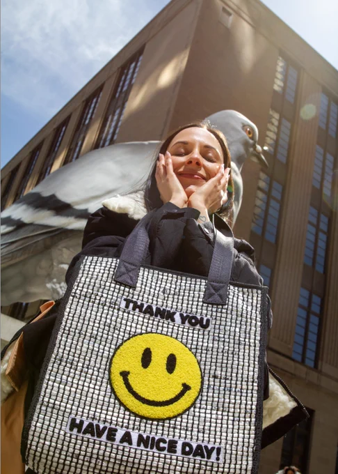 Model Kara Neko poses happily with her ANYBAG in front of New York City's King Pigeon statue, a larger than life homage to the city's unofficial mascot. The bag, made from upcycled plastic, features a smiley face patch that says THANK YOU HAVE A NICE DAY! It's ANYBAG's top seller. Perfect for Sustainable Fashion fans.