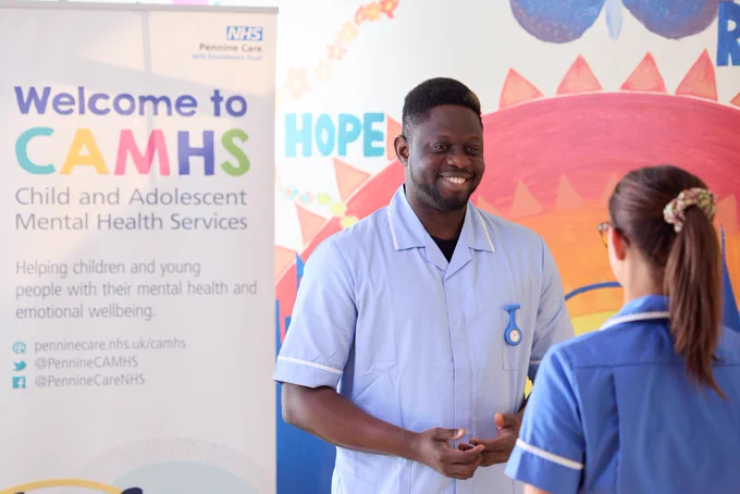 A healthcare professional in a white uniform is smiling and conversing with a colleague in front of a banner reading "Welcome to CAMHS - Child and Adolescent Mental Health Services."