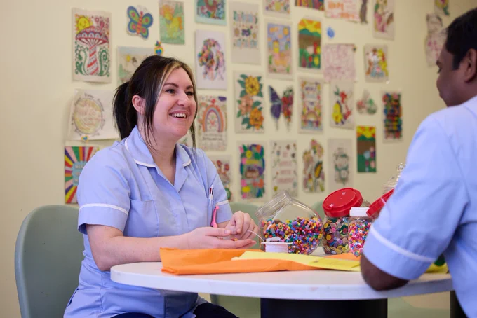 Two healthcare professionals are smiling and conversing at a table, surrounded by colorful craft supplies, in a room decorated with vibrant artwork.