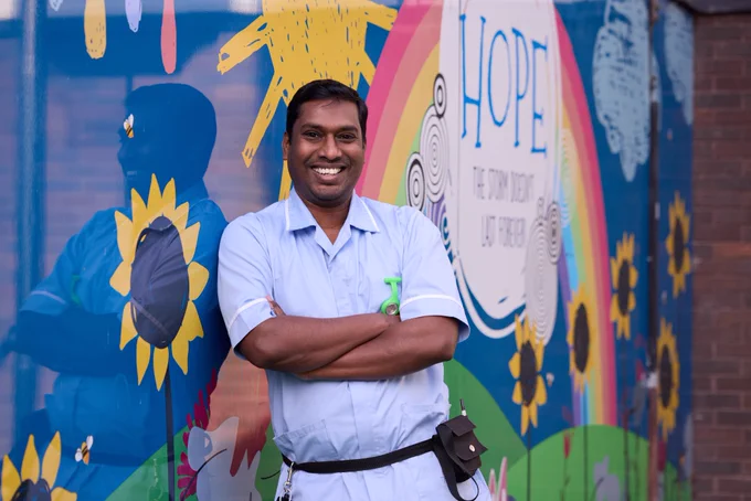 A smiling healthcare worker in scrubs stands in front of a colorful mural with the word "HOPE" prominently displayed.