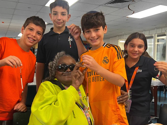 A group photo of 4 students and one senior program participant at Miami Lighthouse during a student volunteer visit. In the photo, they smile for the camera while holding up white cane-shaped keychains that they made together in art class.