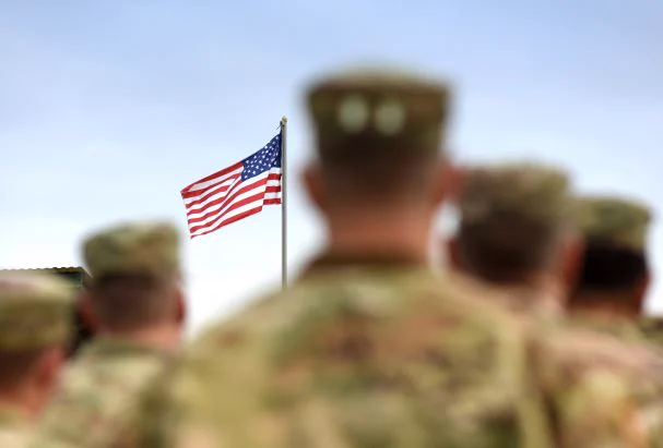 U.S. service members in uniform stand facing a waving American flag, with the flag in focus and the soldiers blurred in the foreground.