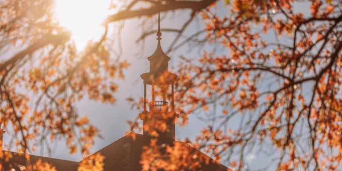 Sunlight filters through autumn leaves, highlighting a lantern atop a classic brick building, creating a warm, picturesque scene.