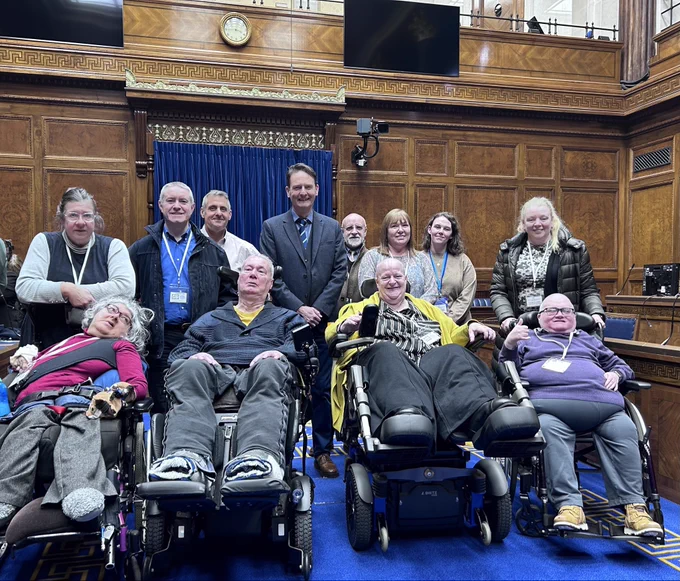 Image of a group of people in the Northern Ireland Assembly Chamber following the Disabled People’s Parliament. All are connected to the Independent Living Fund as recipients, families, employees or civil service.
