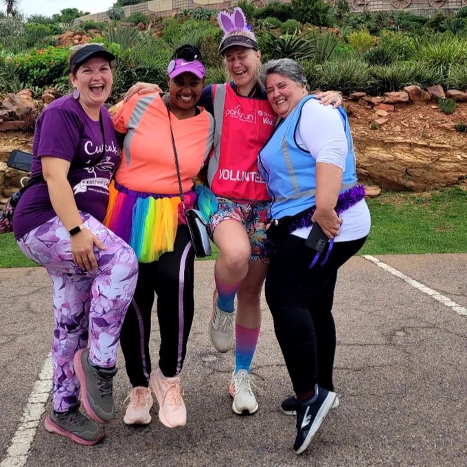 Four parkrunners in colorful outfits smiling joyfully in front of the Voortrekker Monument.
