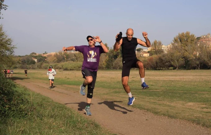 Two parkrunners, one in a purple top and the other in black, joyfully jumping with a running pose in a park.