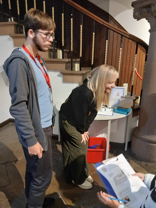 A young man wearing a lanyard and a woman stand by a table and explain a trail sheet to visitors.