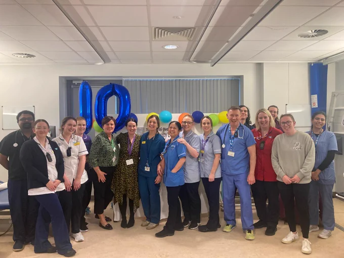 Group of hospital staff wearing uniforms standing in a hospital ward with a balloon shaped in a number 10 in the background