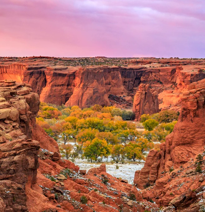 Red sandstone cliffs surround a valley of yellow and green autumn trees with light snow on the canyon floor under a pink sky.