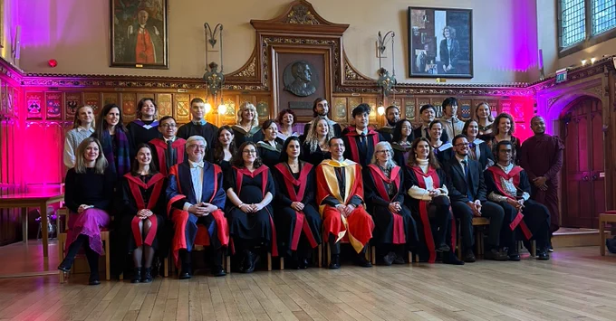Colour photo of School of Divinity graduating students and staff members standing and seated together at the Rainy Hall stage as they pose for a photo