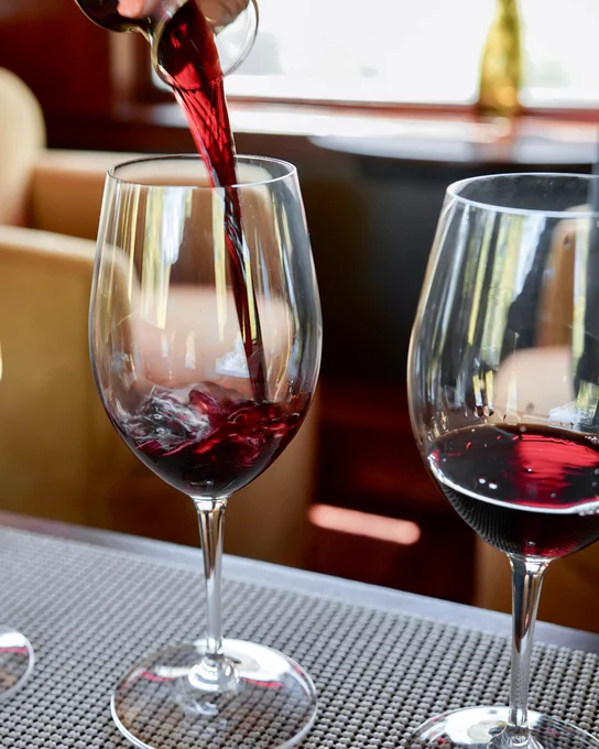 Red wine being poured into a tall glass, with another partially filled glass beside it, set on a textured tablecloth.