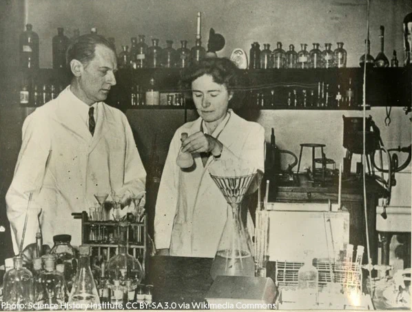 Carl and Gerty Cori, one holding a flask, in a laboratory filled with chemical equipment and bottles on shelves.
