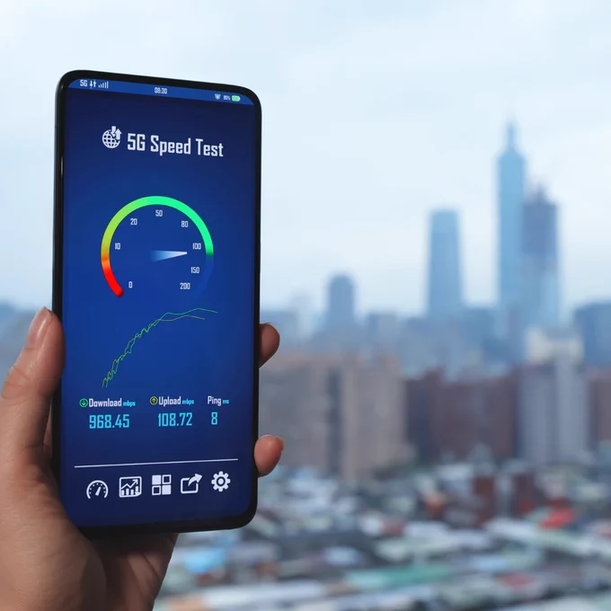 Hand holding a smartphone showing a 5G speed test with download and upload metrics against a blurred city background.