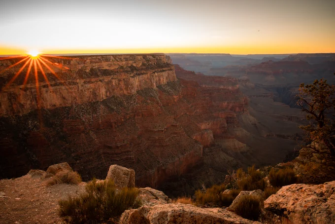 Sunset over the Grand Canyon, casting a warm glow on the layered cliffs. Sparse vegetation in the foreground.