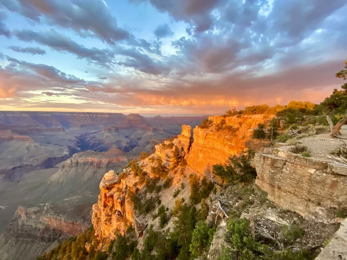 Sunset over the Grand Canyon, lighting up the rock formations with vibrant orange hues. Dramatic clouds enhance the colorful sky.