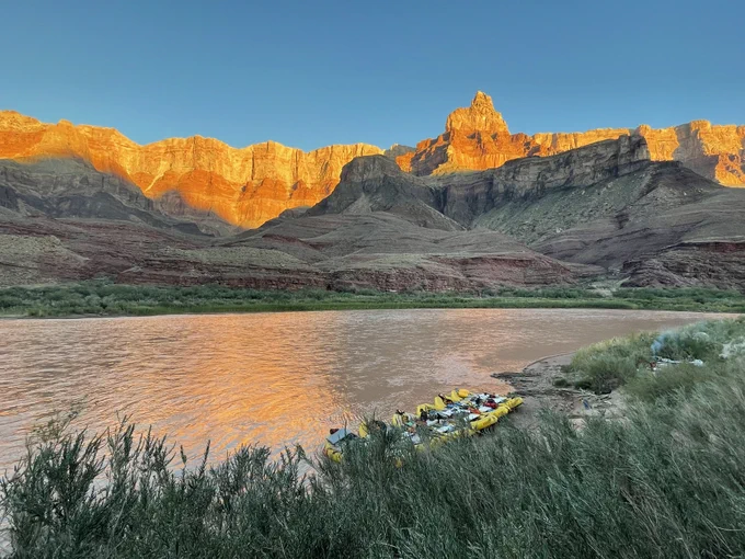 Sunlit Grand Canyon cliffs with a calm river in the foreground, where several yellow rafts are docked on the riverbank.