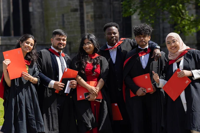 Graduates holding diplomas and smiling, wearing academic regalia outside a stone building.