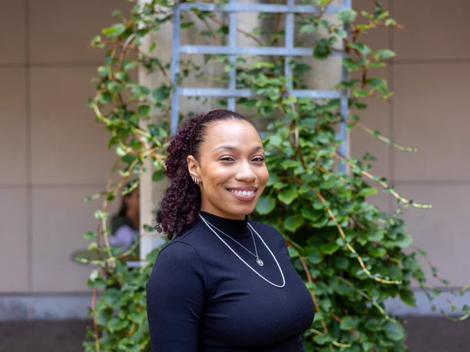 Portrait of a smiling person in front of a vine-covered ladder, wearing a black top and a necklace.