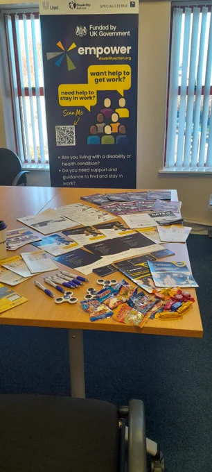 A display table set up for Disability Action. The table is covered with leaflets, brochures, pens, fidget spinners, and sweets. Behind the table is a tall Empower-branded pop-up stand with information about support for disabled people seeking work, including a QR code and the programme logo. The setting appears to be an office room with chairs and vertical blinds.