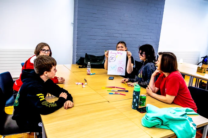 A small group of Young Advisors sit around a table during a workshop. One young person is holding up a colourful sheet of paper covered in drawings and notes, while the others look on and chat. Bright pens, drinks and a hoodie are scattered on the table in the meeting room.