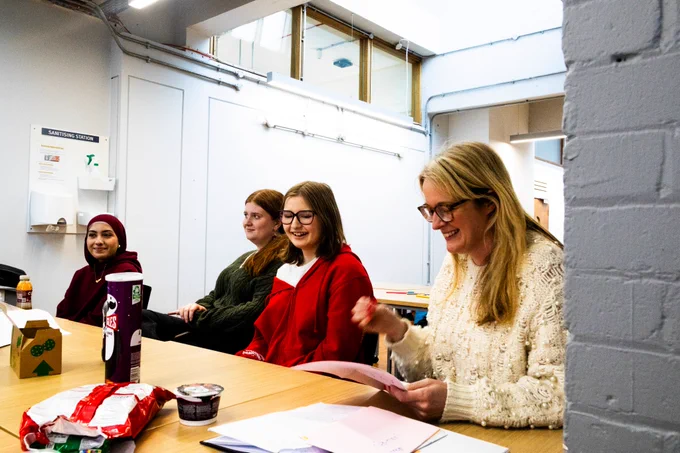 The Commissioner sits at a table with members of the Young Advisors group in a bright meeting room. They are all smiling and laughing as the Commissioner reads from a sheet of paper. Snacks and drinks sit on the table, and a “sanitising station” sign is visible on the wall behind them.