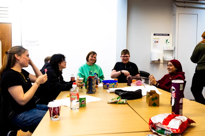 A group of Young Advisors sit together around a table, chatting and laughing during a workshop session. Drinks, snacks and stationery are spread across the table. A sanitising station is visible on the wall behind them in the bright meeting room.
