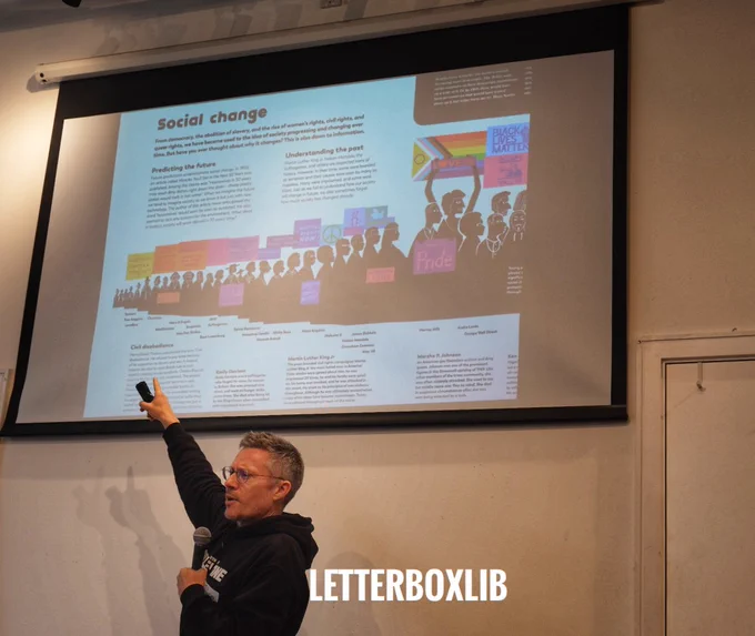 Chris Haughton stands, mic in left hand. Right hand raised, pointing to a slide. This shows an inside double page spread of his book The History of Information.: top title reads Social Change. We see a line of people in silhouette marching, holding placards.