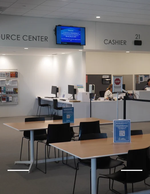 Interior of a Permit Center with tables featuring computers, informational posters and a cashier section marked "21." A person is visible behind the cashier counter.