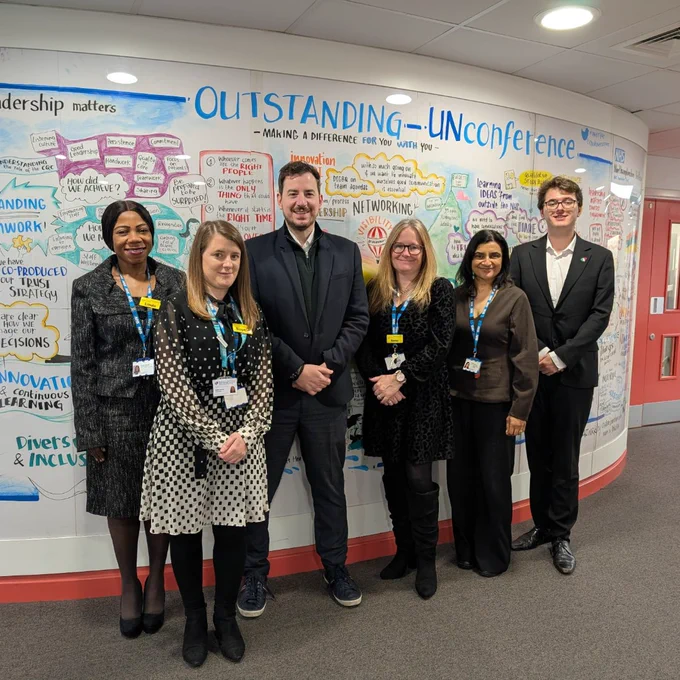 Group of six professionals wearing lanyards smiling in front of a colourful wall with handwritten text on it.
