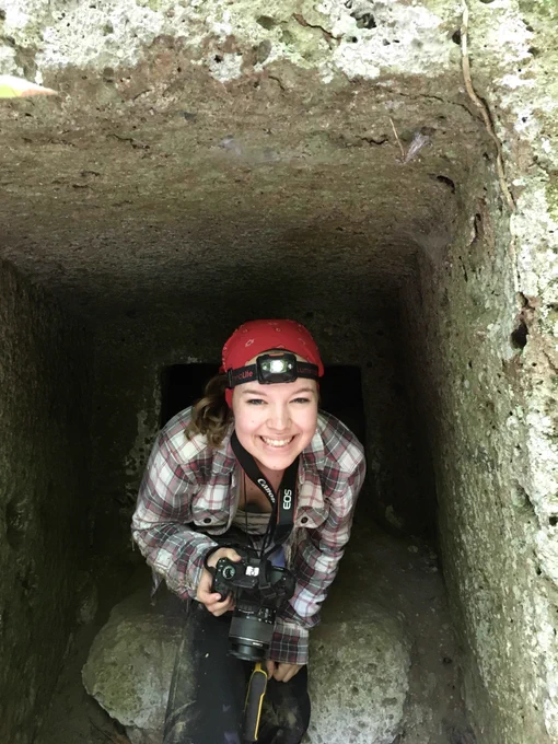Person emerging from a narrow stone tunnel with a camera, wearing a red headlamp and smiling.