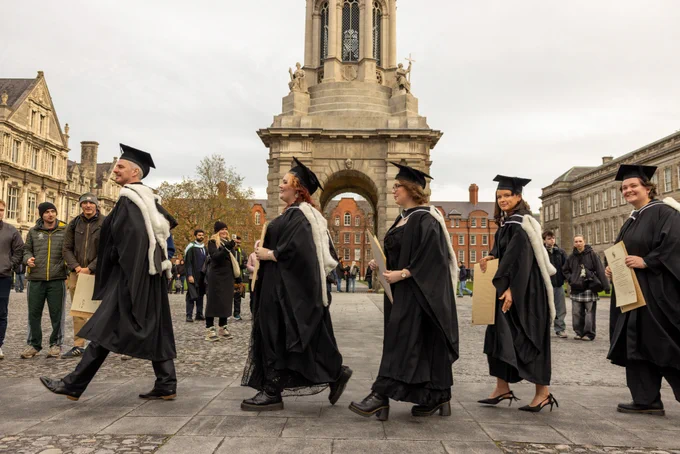 Graduates in academic regalia walking in front of the Front Square at Trinity College Dublin.