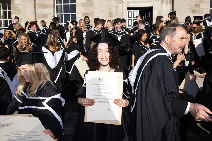 Graduates in caps and gowns celebrating outside, with one person prominently displaying a diploma from the Trinity College Dublin.