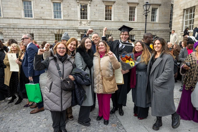 Group of people celebrating a graduation outside a historic building, with one person in a graduation cap and gown holding a bouquet of flowers.