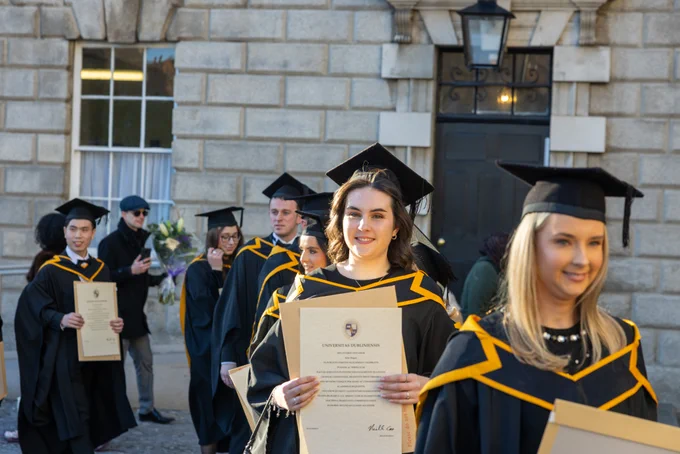 Graduates holding diplomas at a Trinity College Dublin ceremony, walking in front of a historic building.