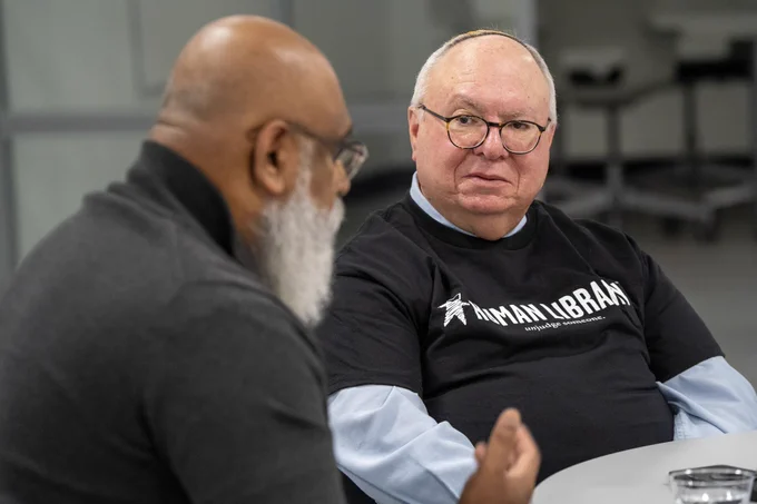 Two individuals engaged in a conversation, one wearing a T-shirt with the "Human Library" logo. They are sitting at a table looking at eachother.