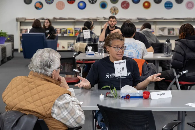 Two people are engaged in conversation at a table in a busy room. In the background, other people are seated at similar tables, and the walls are decorated with colorful round designs.