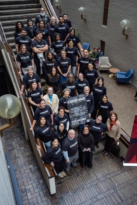 Group of people wearing matching black t-shirts, standing on the stairs inside a building, holding a sign that reads "The Human Library."