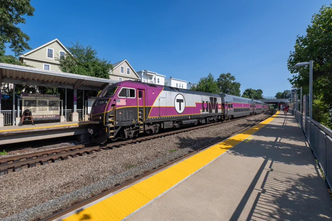 A Commuter Rail train pulling into Morton Street station on the opposite platform on a summer day with blue skies.