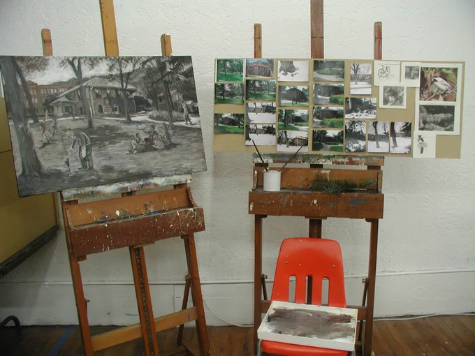 A painting sits on an easel, done in subdued colors that make the scene feel a bid sad. On another easel next to it, someone has pinned multiple photos of the outside of Manitou Springs Library and the surrounding area.