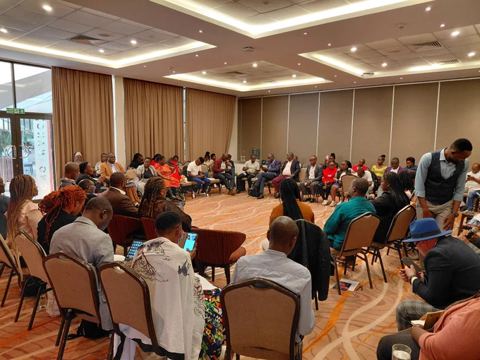 A group of people seated indoors in a circle with a wide gap in the middle.  