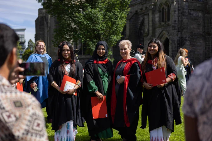 A group of graduates in academic robes and sashes, holding diplomas, smiling during a graduation ceremony on a sunny day with historical stone buildings in the background.