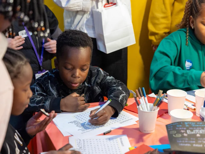 Child at a table engaged in writing or drawing activities, surrounded by others in a lively, colorful workshop setting.