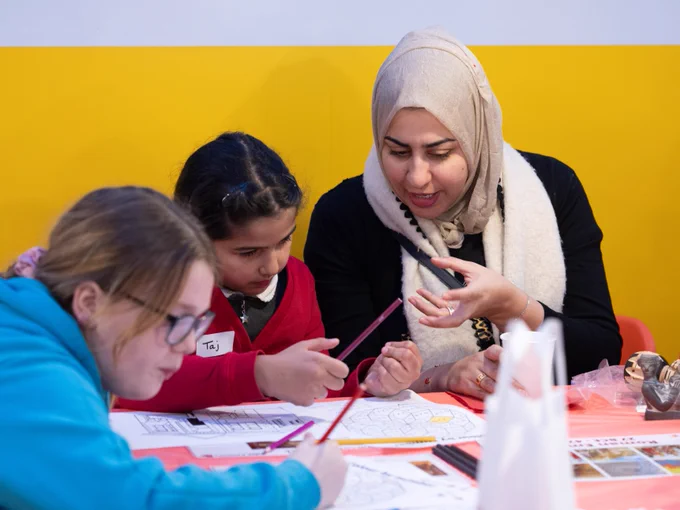 Three individuals engaging in a coloring activity at a table with a yellow background; one adult is instructing two youngsters and papers with various designs are spread out before them.