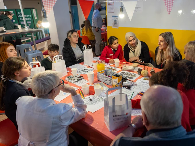 Group of people of diverse ages and backgrounds gathered around tables in a casual event setting, engaging in conversation and activities.