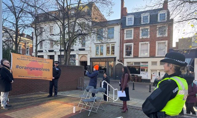 A small outdoor gathering in a town square near a flagpole for an Orange Wolves event. Three individuals stand beside an orange banner that reads ‘Say no to violence against women and girls’ and ‘#orangewolves’. A police officer in a high-visibility vest stands to the right observing the scene.