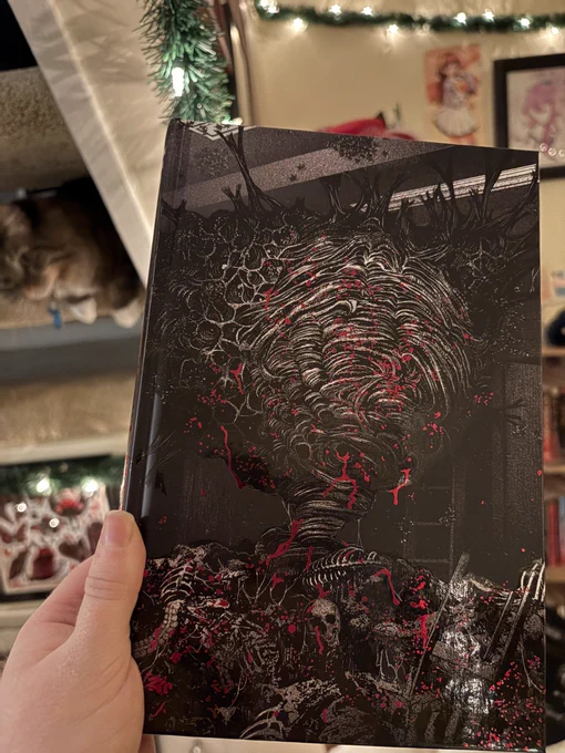 The naked hardback of the book, which is black and white with monochrome red blood splatters. It shows a massive wasp’s nest the size of a room, with skeletons and broken furniture scattered on the floor.