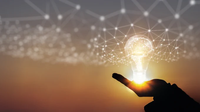 Silhouette of a hand holding a glowing light bulb with a digital brain and network connections inside against a sunset sky.