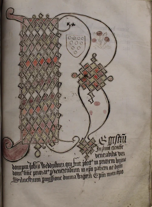 The page has a huge letter R. It is deocrated by a diamond pattern and coloured with maroon, yellow, red, white and pink diamonds. A floral decoration is also in the letter R and a heraldic shield of the Bishops of Worcester. At the base is the name of the Prior whose register it starts.
