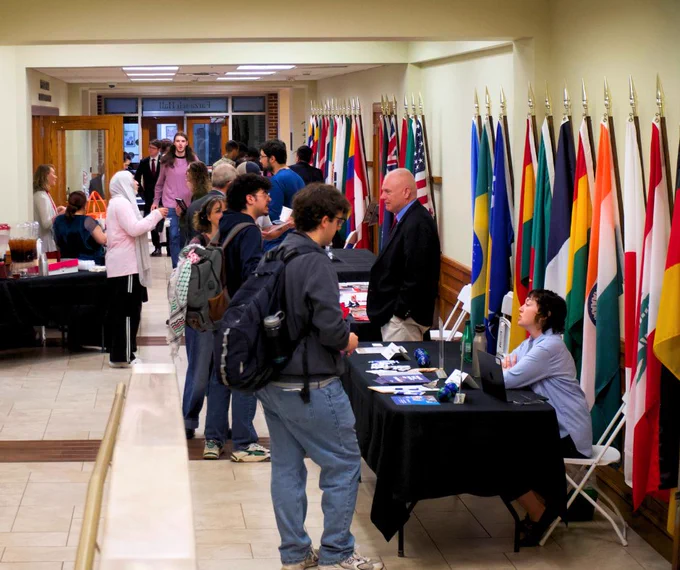 People interact with representatives at various booths in a hall decorated with international flags.